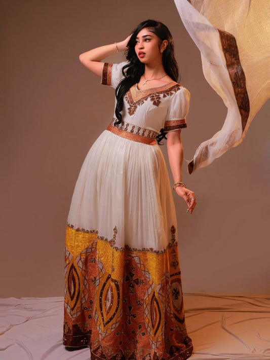 A woman modeling a long white Habesha Dress with brown and gold patterns, featuring short sleeves, a V-neckline, and a wide waist belt, posed against a plain studio backdrop with a flowing sheer fabric.