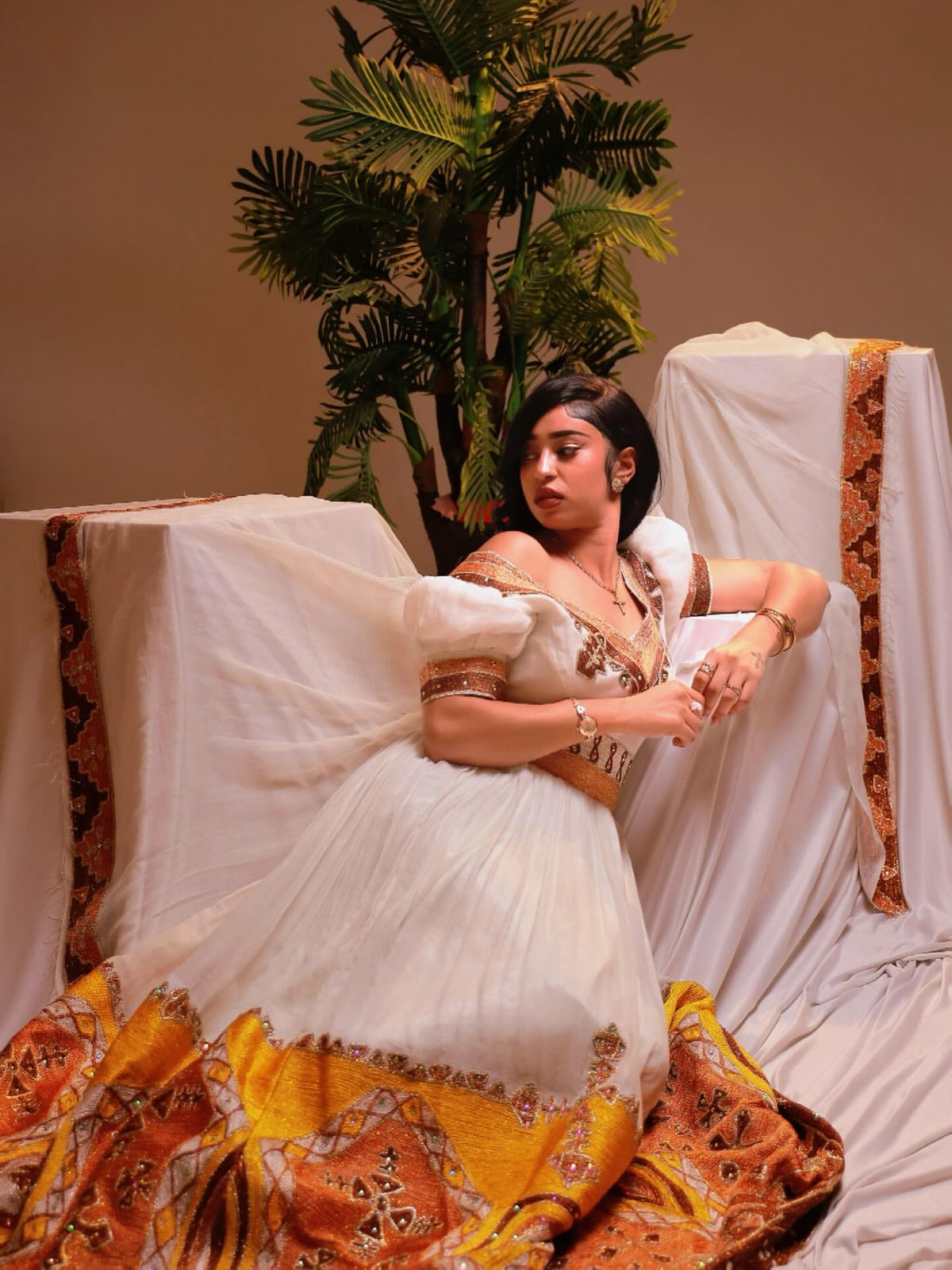 A woman modeling a long white Habesha Dress with brown and gold patterns, featuring short sleeves, a V-neckline, and a wide waist belt, posed against a plain studio backdrop with a flowing sheer fabric.