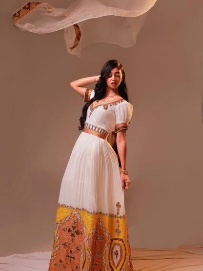 A woman modeling a long white Habesha Dress with brown and gold patterns, featuring short sleeves, a V-neckline, and a wide waist belt, posed against a plain studio backdrop with a flowing sheer fabric.