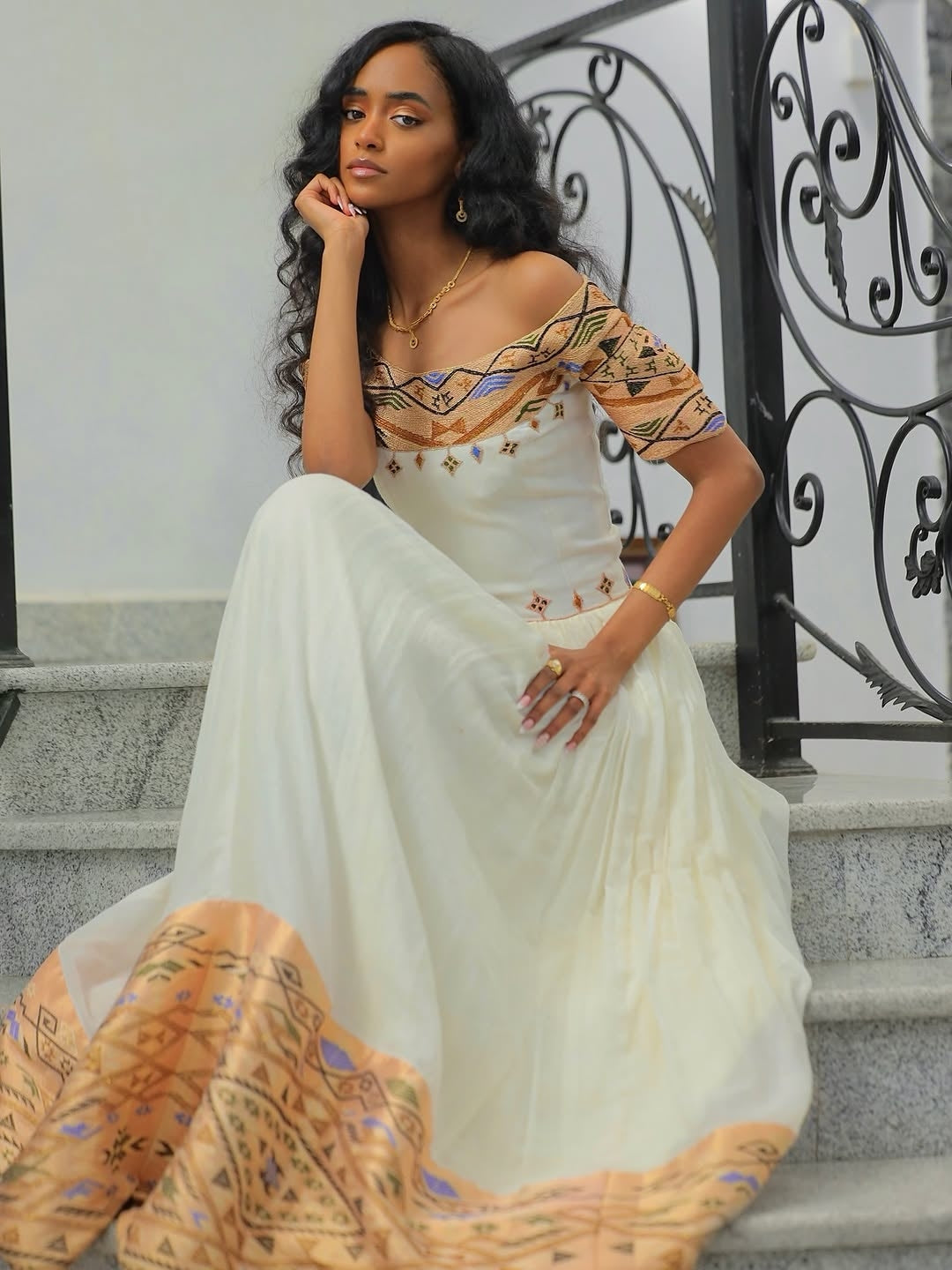 Woman in a white and patterned Gold Habesha Libs sitting on steps with decorative metal railing.