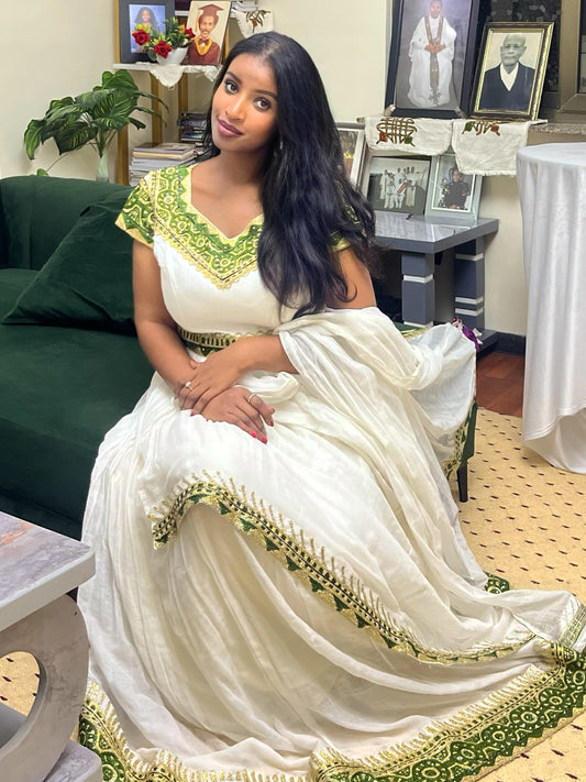 A woman seated indoors wearing a white traditional Ethiopian clothing dress with green embroidered detailing on the neckline, sleeves, and hem. The room includes a dark green sofa, framed photos, a side table, and a white draped table covering.