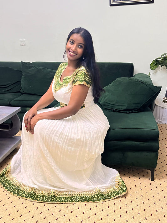 A woman wearing a white pleated traditional Ethiopian clothing with green embroidered details sits on a dark green sofa in a living room setting.