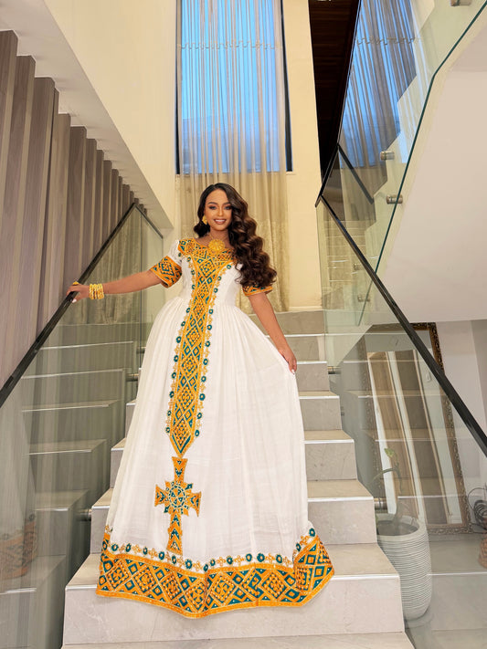 A woman wearing a white traditional Ethiopian Dress with yellow and green embroidery stands on a modern glass-rail staircase indoors.