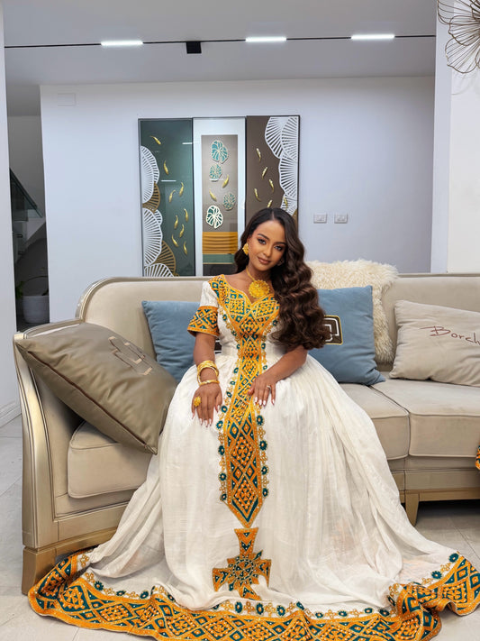 A woman wearing a traditional white Afar Ethiopian Dress with vibrant yellow, green, and blue embroidery, seated on a beige sofa in a modern living room.