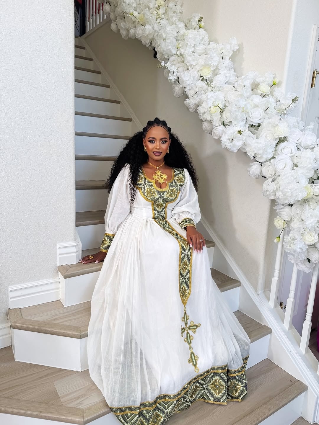 Woman in a white and gold traditional outfit standing on a staircase with floral decorations.