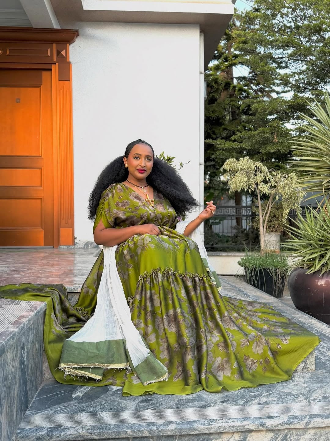 Woman in a green dress sitting outdoors with plants and a building in the background