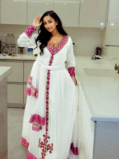 Woman in a white Ethiopian Dress with pink and red patterns standing in a kitchen.