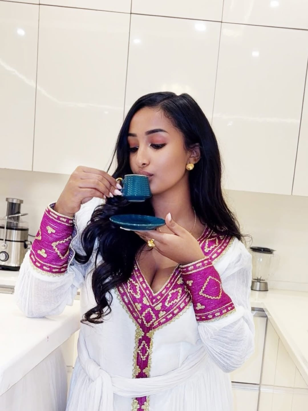 Woman in Ethiopian Dressdrinking from a blue cup in a kitchen.