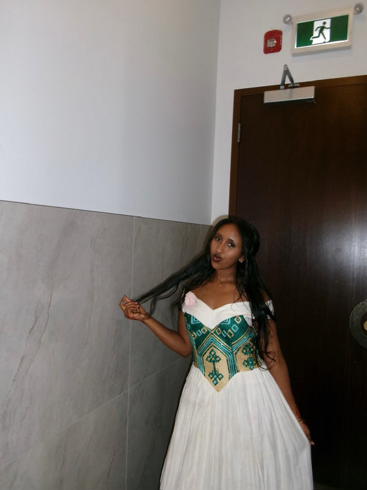 A woman wearing a white A-line  Habesha Kemis  with a green and gold embroidered bodice, standing in an indoor hallway next to a dark wooden door.