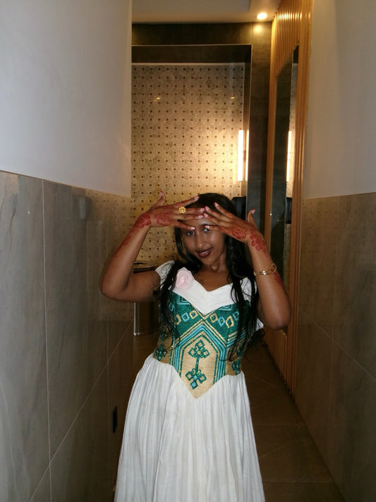 A woman posing in a hallway wearing a traditional Indian  Habesha Kemis  with a white skirt, a green and gold embroidered bodice, and red henna on her hands.