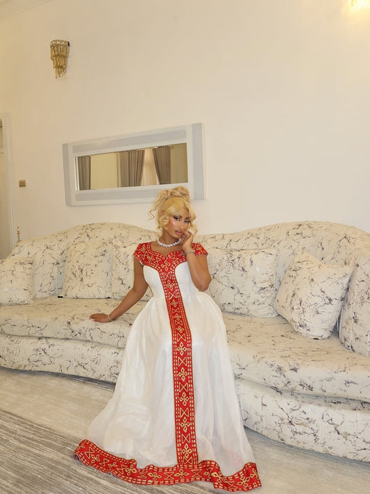 Woman in a white and red habesha kemis sitting on a floral-patterned sofa in a living room.