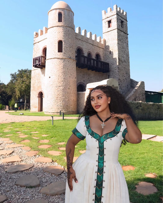 Woman in a white and green habesha dress standing in front of a stone castle.
