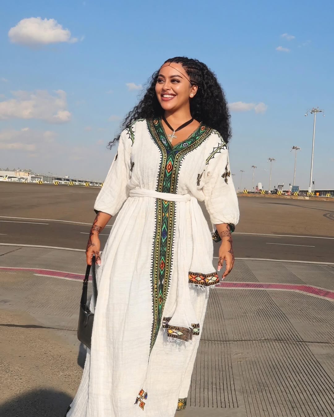 Woman in a habesha dress with green and red patterns standing on an airport tarmac.