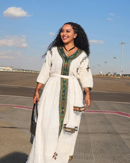 Woman in a habesha dress with green and red patterns standing on an airport tarmac.