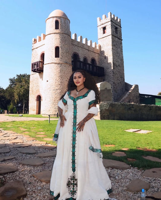 Woman in a habesha dress with green accents standing in front of a stone building with towers.