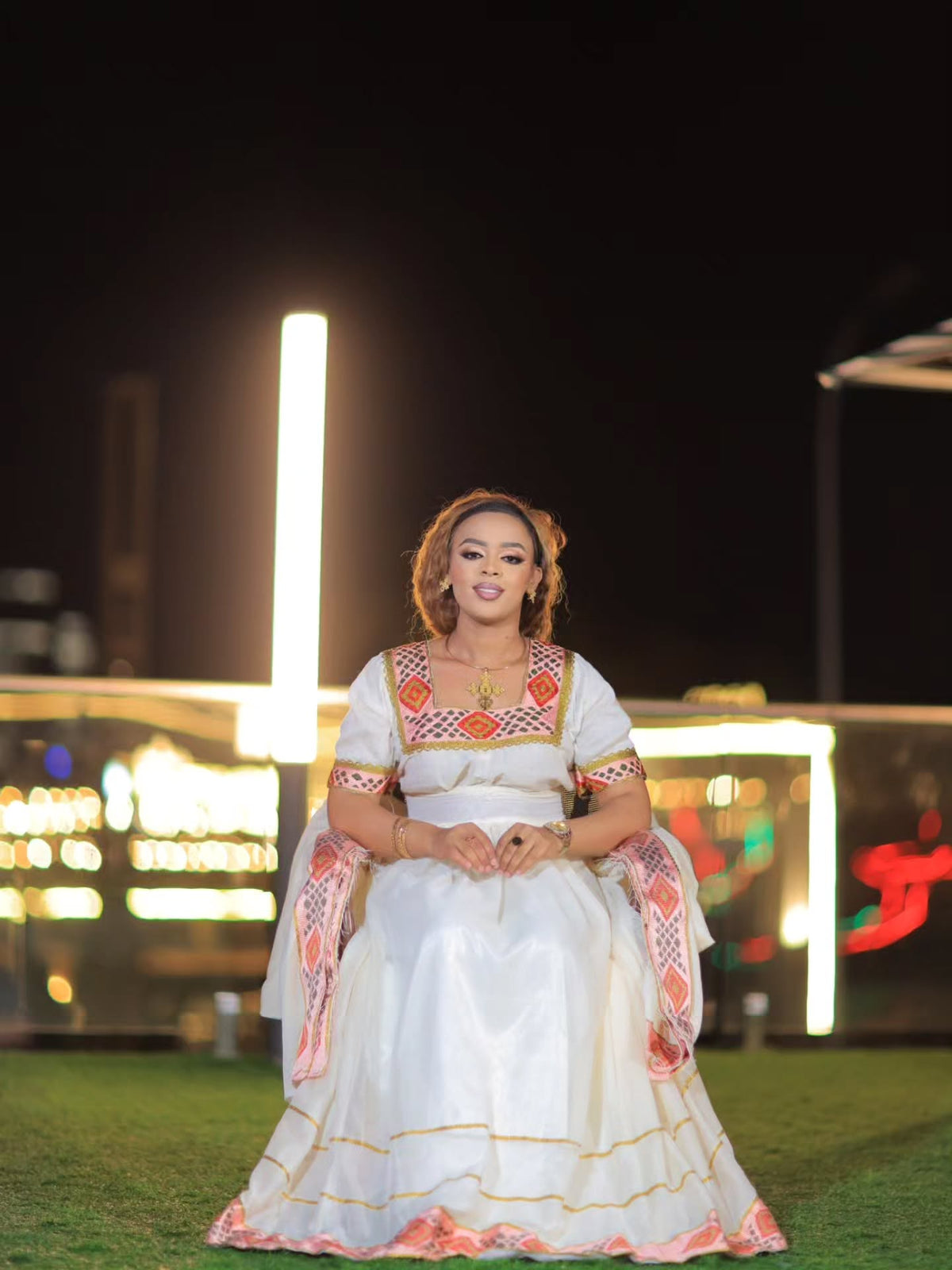 A woman wearing a long white Ethiopian Store Dress with orange and yellow embroidered details stands under a canopy of warm hanging string lights at night.