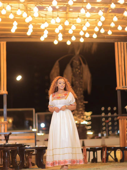 A woman wearing a long white Ethiopian Store Dress with orange and yellow embroidered details stands under a canopy of warm hanging string lights at night.