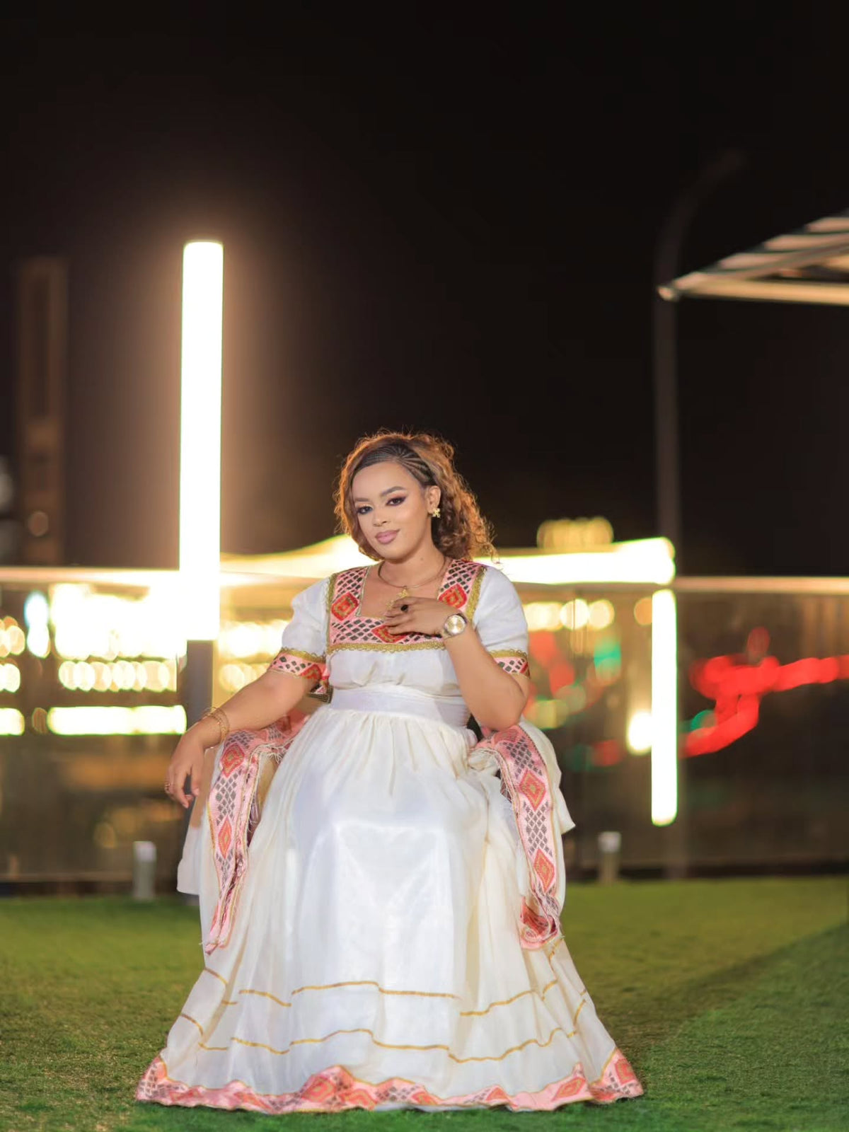 A woman wearing a traditional white embroidered Ethiopian Store Dress with pink and orange accents, standing on artificial grass at night with illuminated background structures.