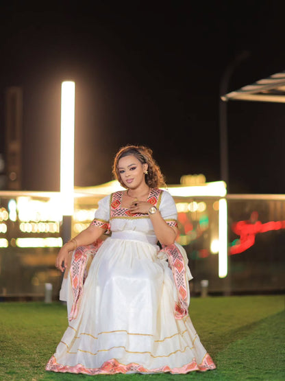 A woman wearing a traditional white embroidered Ethiopian Store Dress with pink and orange accents, standing on artificial grass at night with illuminated background structures.
