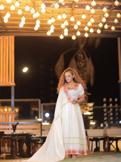 A woman wearing a long white Ethiopian Store Dress with orange and yellow embroidered details stands under a canopy of warm hanging string lights at night.