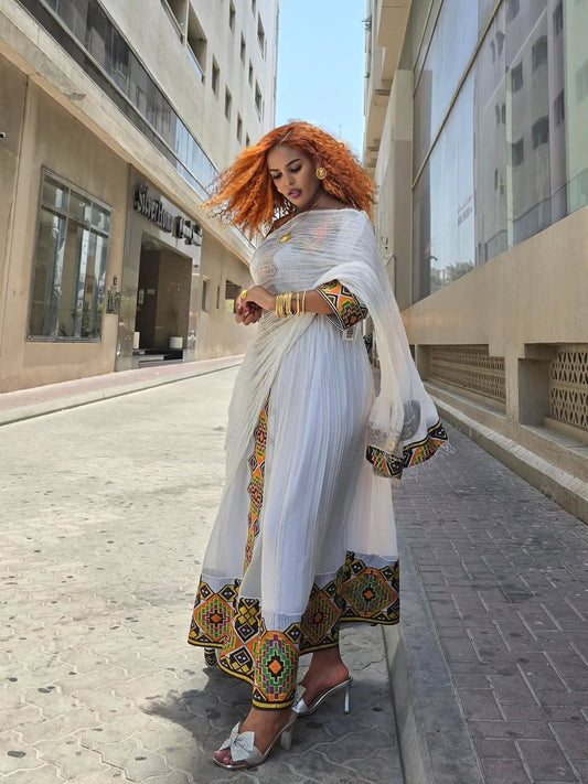 A woman stands on a city street wearing a white traditional habesha zuria  with colorful embroidered borders and a sheer white dupatta. The background shows modern buildings and a paved walkway.