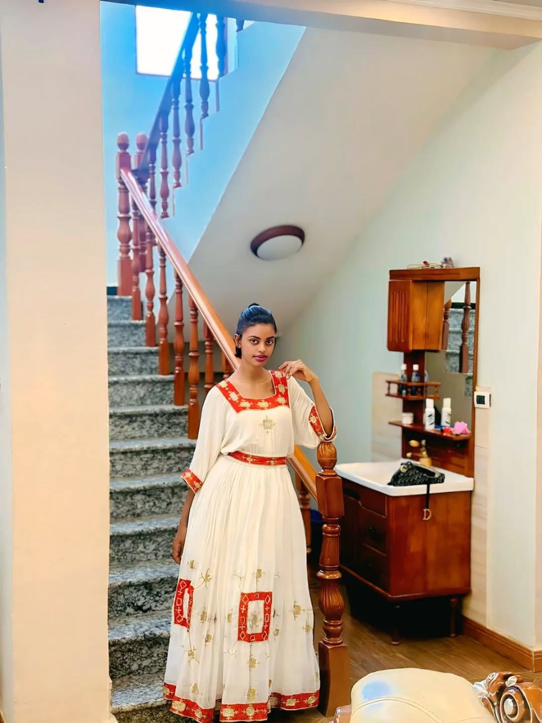 Woman in a white Ethiopian Dress with red patterns standing in a room with wooden furniture and a staircase.