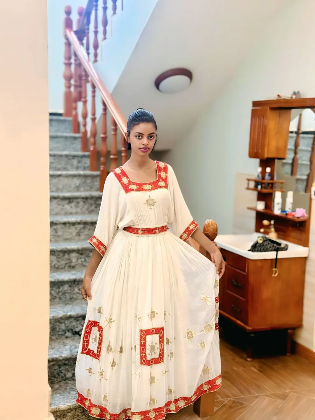 Woman in a white and red Ethiopian Dress standing in a room with wooden furniture and stairs.