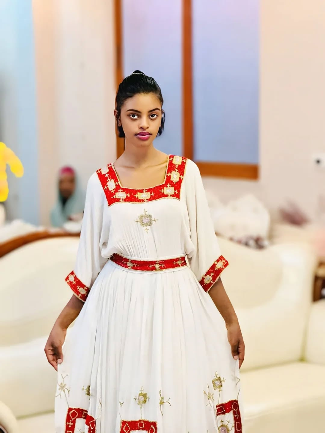 Woman wearing a white Ethiopian Dress with red and gold embroidery in an indoor setting.