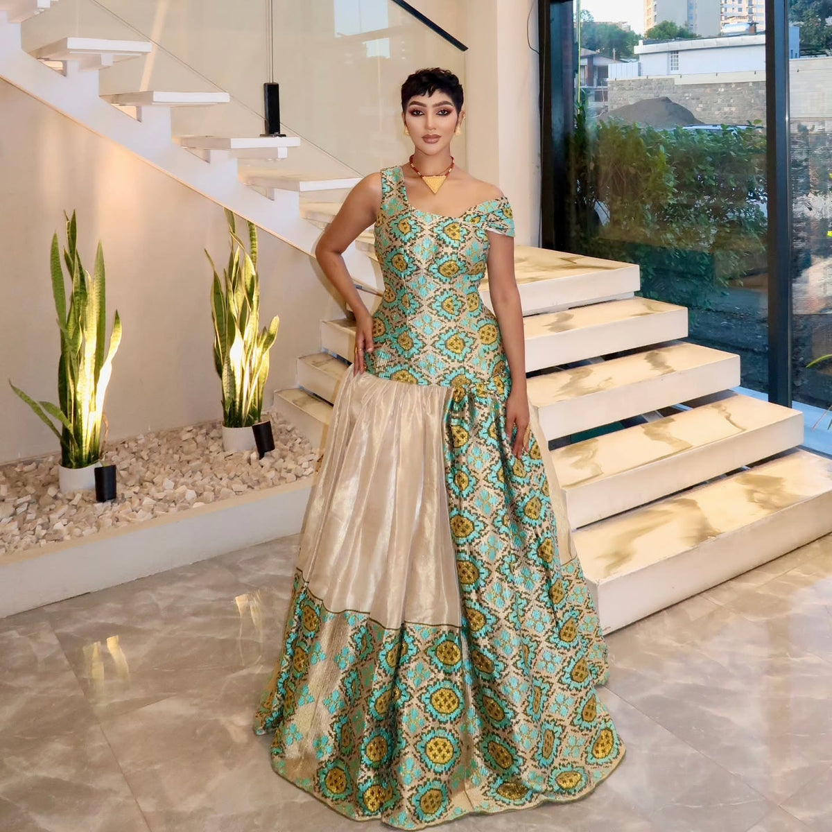 A woman models a floor-length off-the-shoulder Ethiopian Traditional Dress with a teal and gold patterned upper section and a sheer champagne-beige lower skirt, standing in a modern staircase lobby.