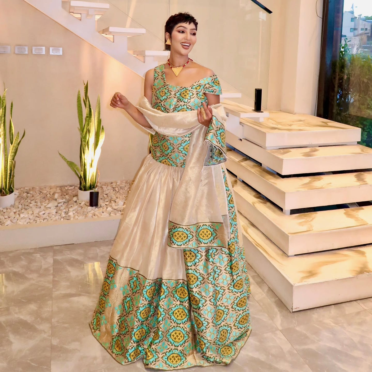 A woman models a traditional Indian lehenga choli Ethiopian Traditional Dress in a modern indoor setting with a staircase. The outfit features a cream-colored skirt with a wide teal and gold patterned border, a matching teal embroidered choli, and a sheer cream dupatta.