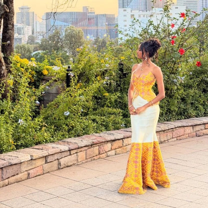 A woman stands outdoors on a paved walkway wearing a strapless yellow and white formal  Habesha Dress  with ornate yellow lace detailing and a flared patterned skirt. Greenery, flowers, and a city skyline are visible in the background.