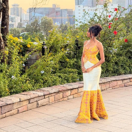A woman stands outdoors on a paved walkway wearing a strapless yellow and white formal  Habesha Dress  with ornate yellow lace detailing and a flared patterned skirt. Greenery, flowers, and a city skyline are visible in the background.