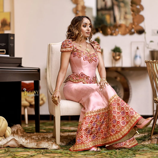 A woman sits on a cream-colored armchair wearing a pink traditional embroidered  Traditional Ethiopian Clothing  with ornate red, gold, and purple patterns on the bodice and flared hem. The setting is an indoor room with a piano, mirror, and decorative elements in the background.