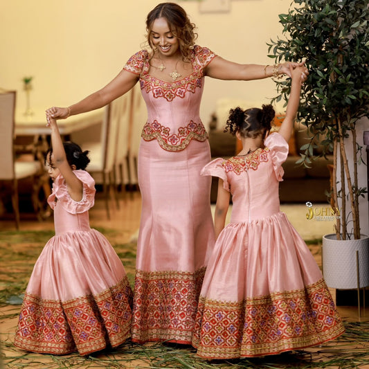 A woman and two young girls wearing matching pink  Traditional Ethiopian Clothing  dresses with ornate red, gold, and multicolor embroidered patterns, posed together indoors with arms raised.