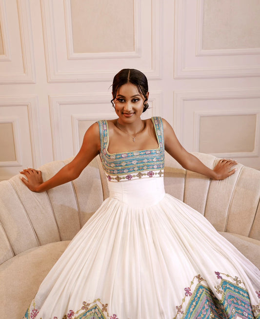 Woman in a white dress with floral embroidery sitting on a beige Ethiopian Dress  against a white paneled wall.