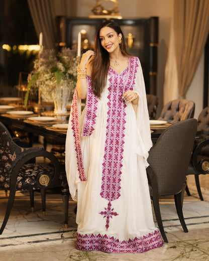 A woman models a white traditional long Habesha Kemis with vibrant magenta embroidery and a matching dupatta in an elegant, warmly lit dining room setting.