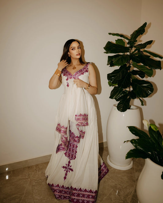 A woman wearing a white sleeveless maxi Habesha Kemis with purple and magenta embroidery, standing in an indoor setting next to large potted green plants.