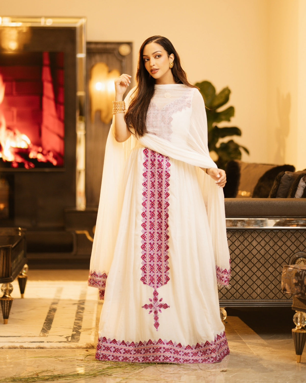 A woman modeling a traditional white embroidered Habesha Kemis with a matching dupatta in a warm, elegant indoor setting with a fireplace in the background.