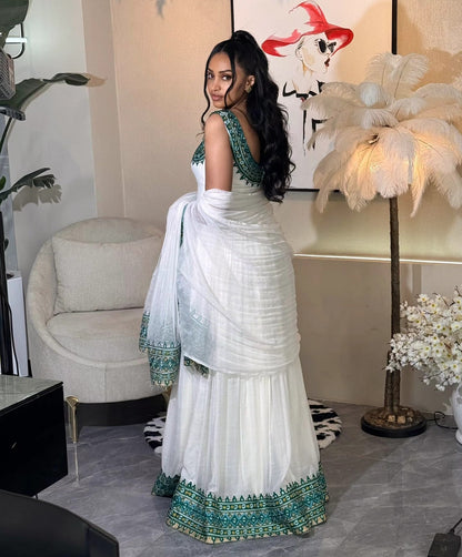 A woman wearing a white traditional Traditional Ethiopian Clothing with green embroidered details, standing in a decorated indoor room with a chair, lamp, and wall art.
