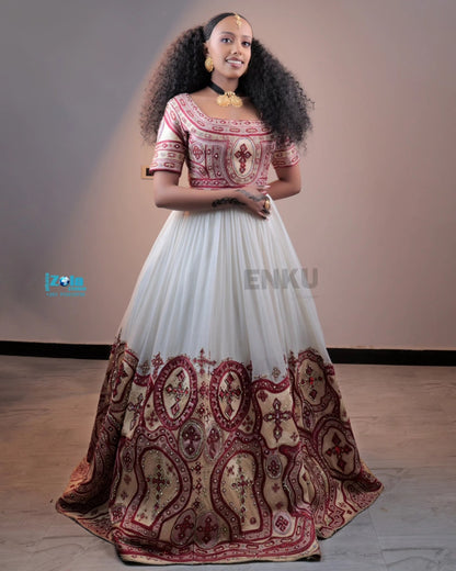 A woman modeling a traditional Ethiopian full-length Habesha matching couple outfit with a white tulle skirt featuring a large maroon paisley border, and a short-sleeved bodice with maroon and gold embroidery.