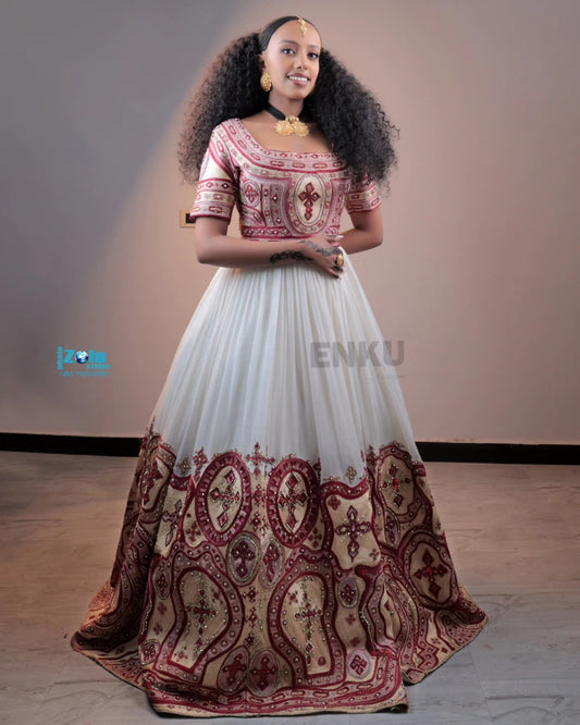 A woman modeling a traditional Ethiopian full-length Habesha matching couple outfit with a white tulle skirt featuring a large maroon paisley border, and a short-sleeved bodice with maroon and gold embroidery.