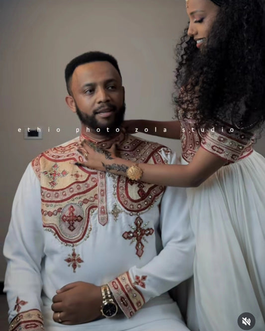 A man wearing a white traditional embroidered tunic with red and gold Habesha matching couple outfit sits while a woman in a matching white dress adjusts his collar. The photo has a watermark reading 'ethio photo zola studio' across the center.