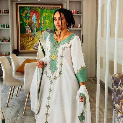 A woman wearing a white kurta with green and gold floral Traditional Ethiopian Clothing, standing in a modern dining room setting.