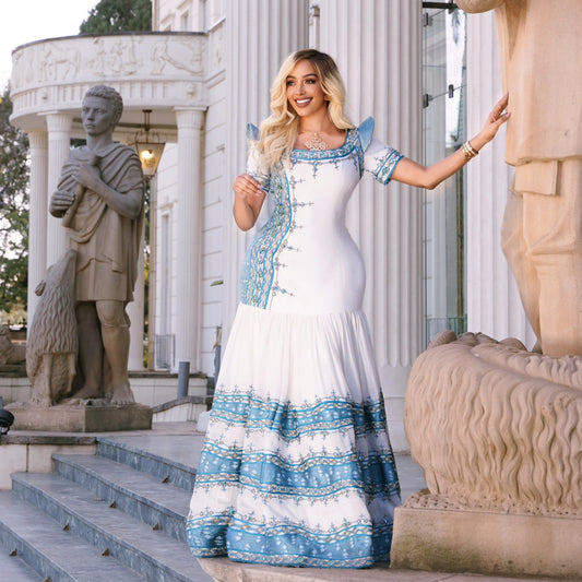 A woman wearing a white and blue patterned Traditional Ethiopian Clothing stands on stone steps in front of a classical building with statues.