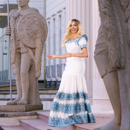 A woman stands outdoors between large stone statues wearing a white maxi Traditional Ethiopian Clothing with blue patterned trim and a blue-and-white patterned hem. The background shows a classical building facade with columns and steps.