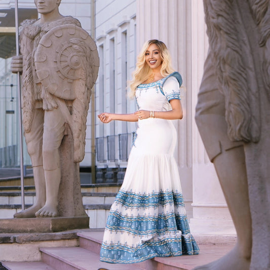 A woman stands outdoors between large stone statues wearing a white maxi Traditional Ethiopian Clothing with blue patterned trim and a blue-and-white patterned hem. The background shows a classical building facade with columns and steps.