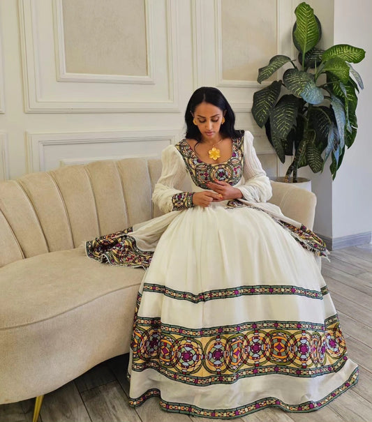 A woman sits on a beige sofa wearing a traditional white embroidered Ethiopian Traditional Dress with colorful floral patterns and a gold necklace, in an indoor room with a potted plant.