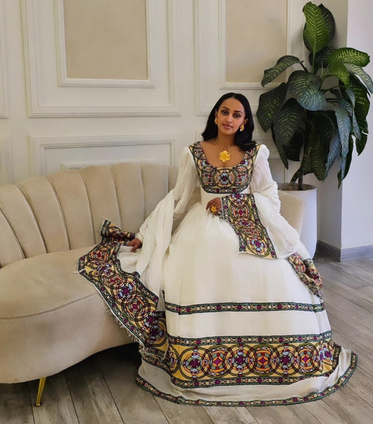 A woman wearing a traditional white Ethiopian Traditional Dress with colorful embroidered borders sits on a beige sofa in a living room setting.