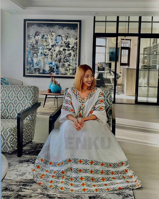 A woman wearing a white A-line maxi Ethiopian Dress with colorful geometric embroidery sits in a modern living room setting.