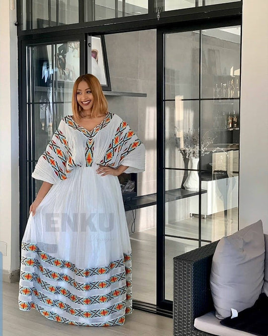 A woman wearing a white kaftan Ethiopian Dress with colorful geometric embroidery, standing in front of a glass sliding door in a modern interior.